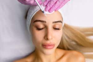 Close-up of a young cosmetologist’s hands injecting Botox into a woman’s face while smiling, as the woman relaxes with her eyes closed