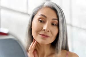 Portrait of happy middle-aged woman touching face in mirror, concept of lifting, anti-wrinkle skincare, and spa beauty treatments.