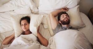 Frustrated woman covering ears in bed beside snoring man, concept of sleep apnea, loud snoring, and disturbed rest at home.
